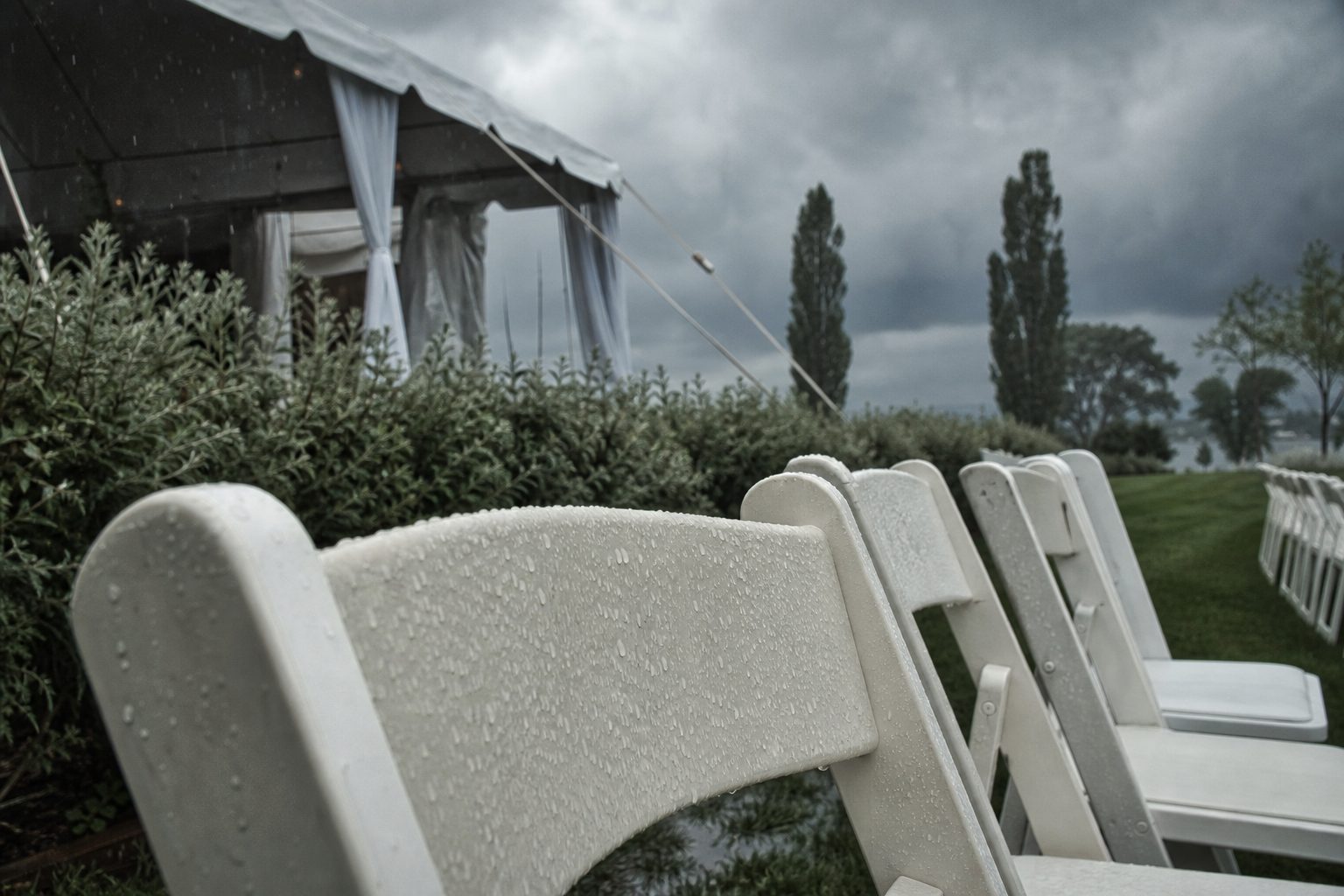 White folding chairs on a rain soaked lawn beside an open tent under a heavy gray sky